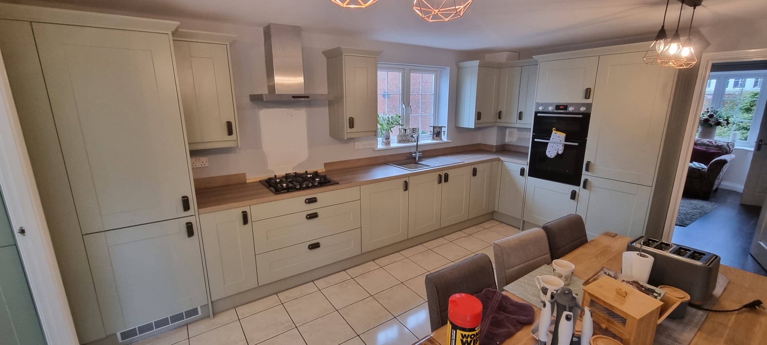 Sage green shaker kitchen with oak-effect worktops, bronze handles and integrated double oven by LMB Joinery.